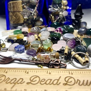 May include: A collection of gemstones, charms, and decorative items. The assortment includes various colorful stones, small glass bottles, and metal charms. A decorative fork and a wooden ruler with the text "Drop Dead Druzy" are also present.