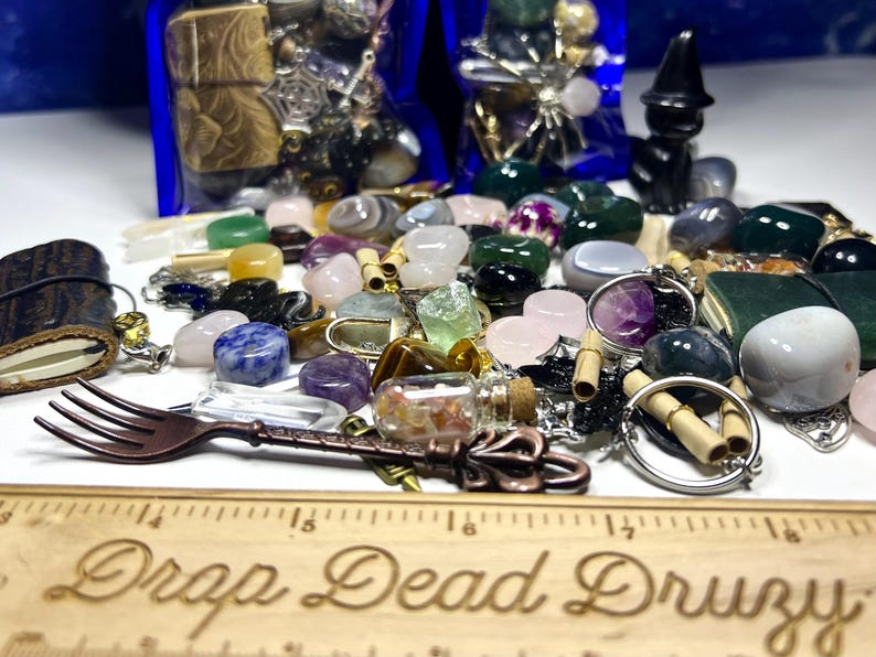 May include: A collection of gemstones, charms, and decorative items. The assortment includes various colorful stones, small glass bottles, and metal charms. A decorative fork and a wooden ruler with the text "Drop Dead Druzy" are also present.
