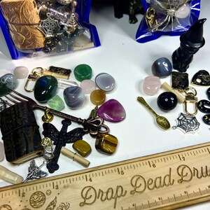 May include: Assortment of crystals, charms, and decorative items. Includes a wooden ruler with "Drop Dead Druzy" text, a black cat figurine, a golden spoon, and various gemstones in shades of green, pink, and purple.