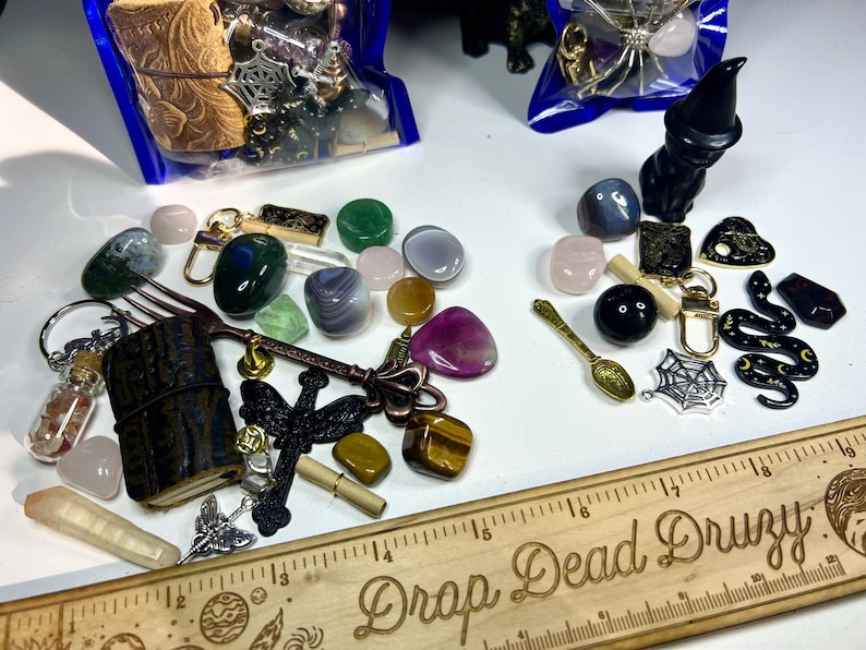 May include: Assortment of crystals, charms, and decorative items. Includes a wooden ruler with "Drop Dead Druzy" text, a black cat figurine, a golden spoon, and various gemstones in shades of green, pink, and purple.