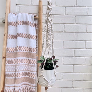 May include: A cream macrame plant hanger with a white ceramic pot holding a green and burgundy plant. The hanger is suspended from a wooden ring against a white brick wall. A wooden ladder and a white and beige towel are in the background.
