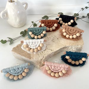 May include: Assortment of handmade macrame wall hangings with wooden beads and fringe. The hangings are in various colours, including blue, green, brown, pink, and black.