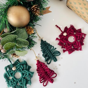 May include: Two macrame Christmas tree ornaments in dark green and deep red, each with a gold star topper, and two macrame snowflake ornaments in dark green and deep red, each featuring a small wooden bead, arranged on a white surface.