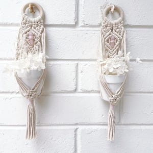 May include: Two macrame plant hangers with white ceramic pots filled with white flowers. Each hanger has a wooden ring, a pink crystal, and a tassel. The hangers are displayed against a white brick wall.