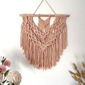 May include: A blush-coloured macrame wall hanging. The handmade textile art piece features intricate knotting patterns and a fringe. It is suspended from a wooden dowel and hangs against a white wall. Decorative flowers are in the foreground.