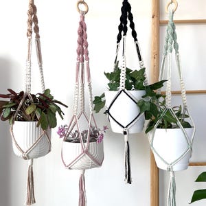 May include: Four macramé plant hangers in different colours, each holding a white ceramic pot with greenery. The hangers are suspended from a wooden rod, showcasing a bohemian style with natural and pastel tones. Each hanger has a tassel.