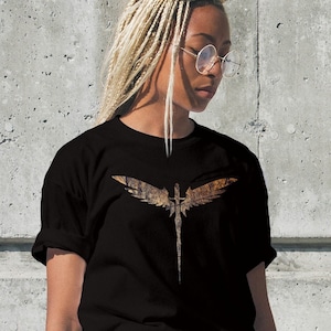 May include: A black short-sleeve t-shirt featuring a central graphic print of a sword with large, textured, brown and gold wings extending from the hilt. The design has a weathered, distressed appearance, contrasting against the solid black fabric.