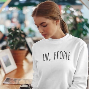 May include: White sweatshirt with the text "EW, PEOPLE" in black. The long-sleeved crew neck sweatshirt is worn by a person holding a floral-patterned coffee cup. The person is in a cafe setting.