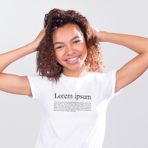 Puede incluir: Camiseta blanca con el texto "Lorem ipsum" en negro. La camiseta la lleva una persona con el pelo castaño rizado, sonriendo y con las manos cerca de la cabeza. La persona lleva pantalones negros.