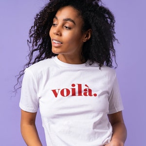 May include: White t-shirt with the word "voilà" in red lettering. The shirt is short-sleeved and worn by a person with curly dark hair and blue jeans. The background is a solid purple color.