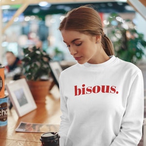 Peut inclure: Un sweat-shirt blanc avec le mot "bisous" en lettres rouges. Le sweat-shirt est à manches longues et a un col rond. Une tasse de café noire avec un motif floral est tenue dans la main.