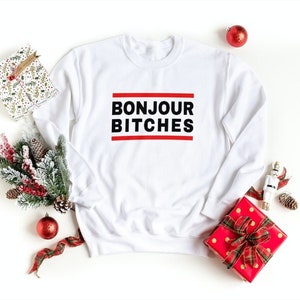 May include: White crewneck sweatshirt with the text "BONJOUR BITCHES" in black, framed by red lines. The sweatshirt is surrounded by holiday decorations, including wrapped gifts, ornaments, and greenery.