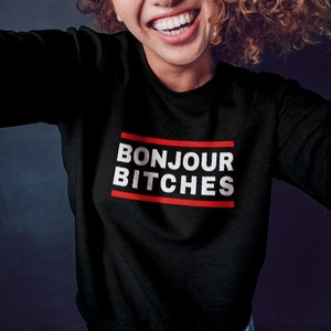 May include: Black sweatshirt with the text "BONJOUR BITCHES" in white, inside a red-lined rectangle. The sweatshirt is being worn by a person with curly hair, smiling at the camera.