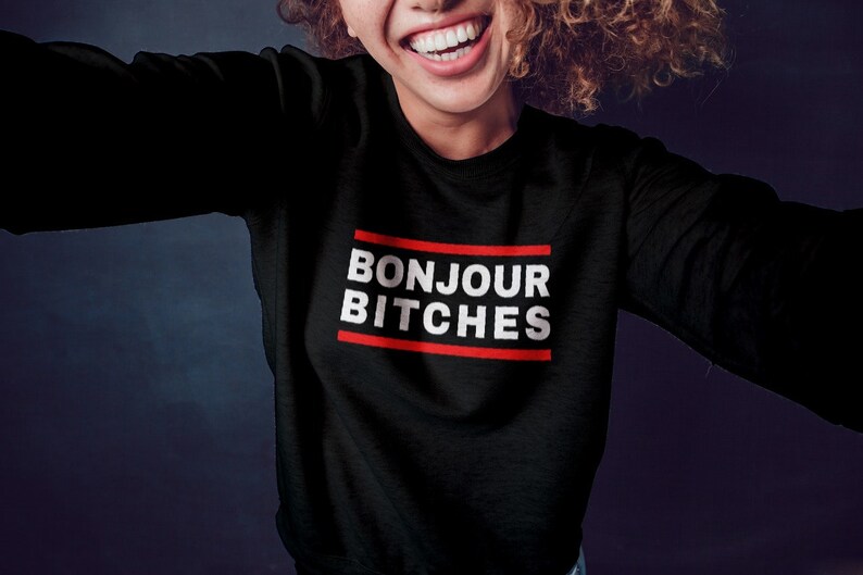 May include: Black sweatshirt with the text "BONJOUR BITCHES" in white, inside a red-lined rectangle. The sweatshirt is being worn by a person with curly hair, smiling at the camera.