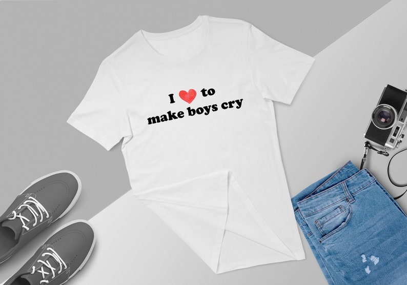 Puede incluir: Camiseta blanca con el texto "I ❤️ to make boys cry" en fuente negra y un coraz&oacute;n rojo. La camiseta se muestra con zapatillas grises, vaqueros azules y una c&aacute;mara vintage.