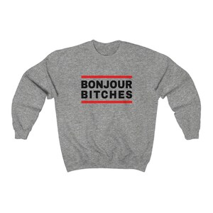 May include: A heather gray sweatshirt with the phrase "BONJOUR BITCHES" in black block letters, framed by two red horizontal lines. The sweatshirt has long sleeves and a crew neck.