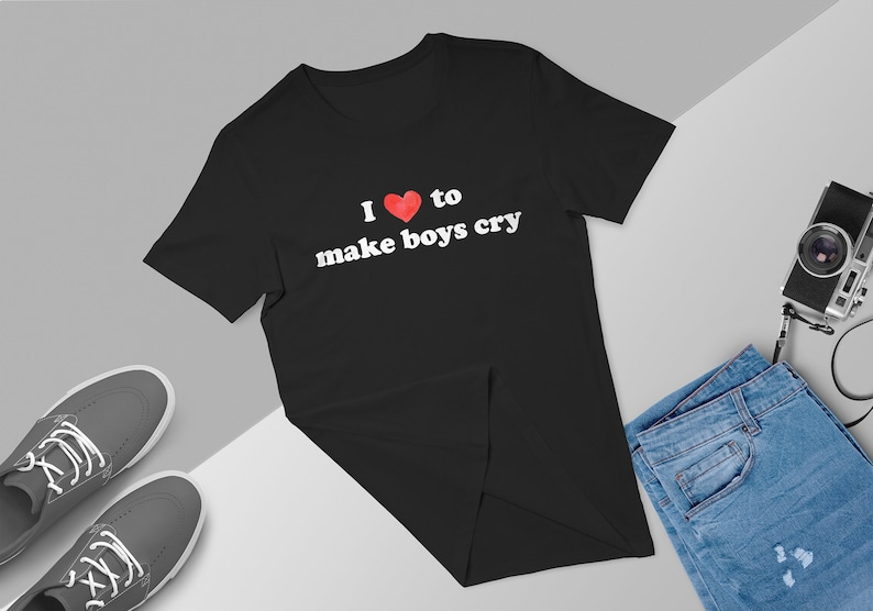 Puede incluir: Camiseta negra con el texto "I ❤️ to make boys cry" en letras blancas y un coraz&oacute;n rojo. La camiseta se muestra sobre una superficie gris y blanca con un par de zapatillas grises, unos vaqueros azules y una c&aacute;mara.