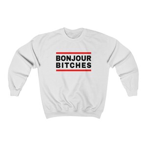 May include: White sweatshirt with the text "BONJOUR BITCHES" in black, framed by two red horizontal lines. The sweatshirt is a casual, long-sleeved garment.
