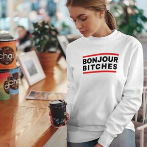 May include: White sweatshirt with the text "BONJOUR BITCHES" in black letters with a red line above and below the text. The model is holding a black coffee cup with a floral design.