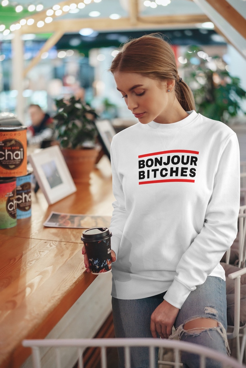 May include: White sweatshirt with the text "BONJOUR BITCHES" in black letters with a red line above and below the text. The model is holding a black coffee cup with a floral design.
