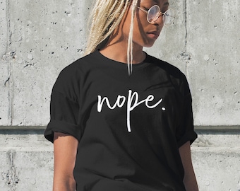 May include: Black t-shirt with the word "nope" in white script. The shirt is worn by a person with blonde braids and round glasses, set against a gray concrete wall.