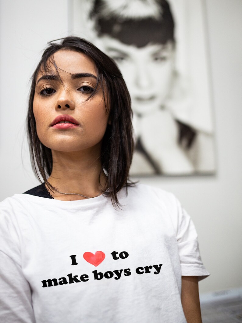 Puede incluir: Una camiseta blanca con la frase "I love to make boys cry" impresa en negro, con un coraz&oacute;n rojo reemplazando la palabra "love". Una mujer con cabello oscuro y maquillaje lleva la camiseta. Un retrato en blanco y negro cuelga en el fondo.