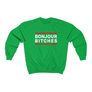 May include: A green sweatshirt with the text "BONJOUR BITCHES" in white, framed by red lines. The sweatshirt is a solid green color, and the text is centered on the chest.