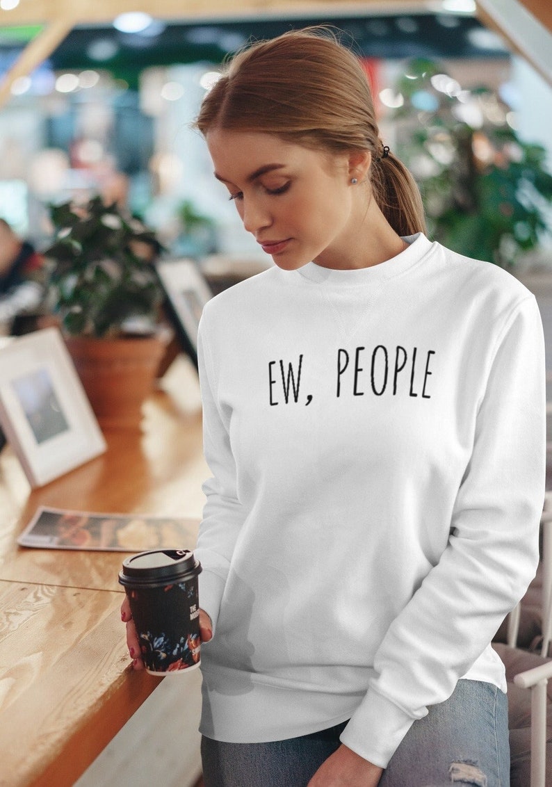 May include: White sweatshirt with the text "EW, PEOPLE" in black. The long-sleeved crew neck sweatshirt is worn by a person holding a floral-patterned coffee cup. The person is in a cafe setting.