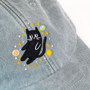 May include: A gray baseball cap with an embroidered black cat surrounded by stars, planets, and moons. The cat is looking up with a playful expression.