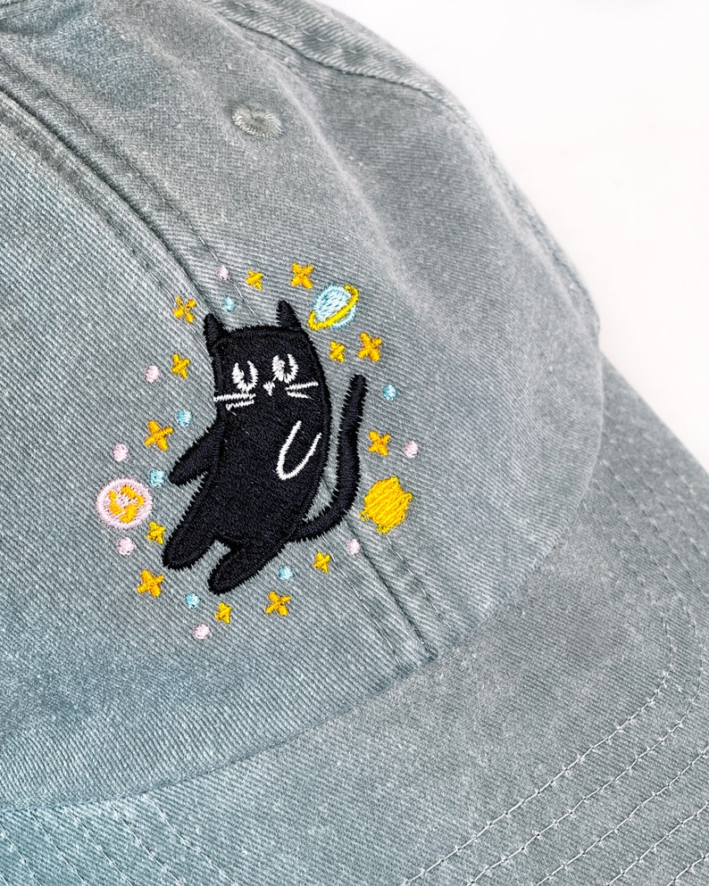 May include: A gray baseball cap with an embroidered black cat surrounded by stars, planets, and moons. The cat is looking up with a playful expression.