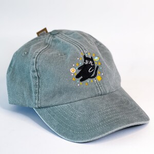 May include: A green baseball cap with an embroidered black cat surrounded by stars and planets.