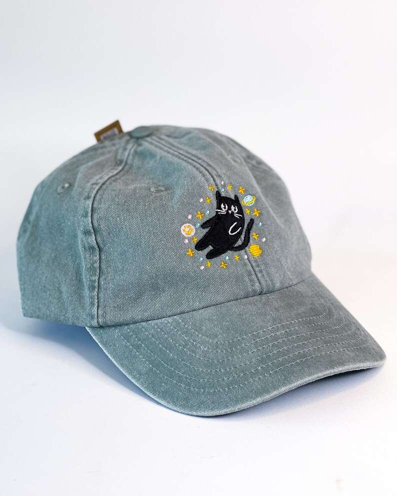 May include: A green baseball cap with an embroidered black cat surrounded by stars and planets.
