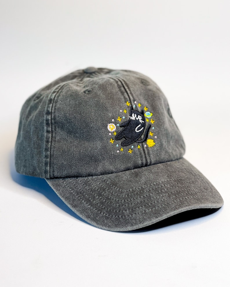 May include: A black and gray baseball cap with an embroidered design of a black cat in space with yellow stars and planets.