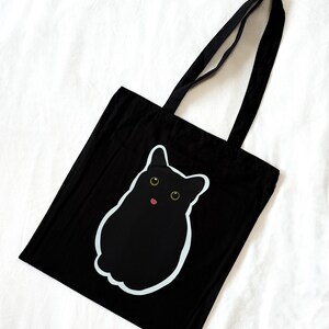 Puede incluir: Un bolso tote negro con una ilustración gráfica de un gato negro sólido con grandes ojos amarillos y una pequeña lengua rosa. El diseño del gato está delineado con un borde blanco fino que contrasta con la tela oscura del bolso.