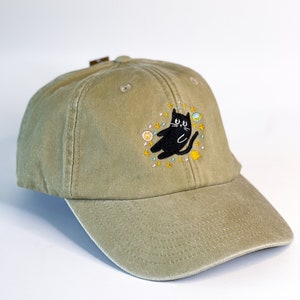 May include: A khaki green baseball cap with an embroidered black cat surrounded by stars and planets.