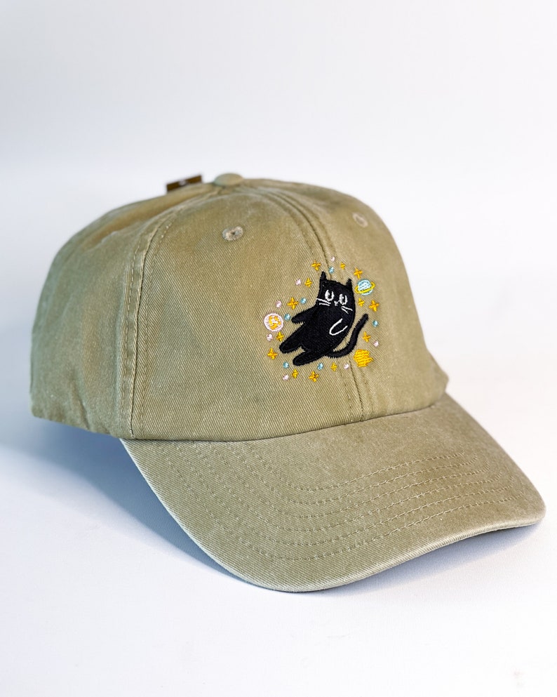 May include: A khaki green baseball cap with an embroidered black cat surrounded by stars and planets.