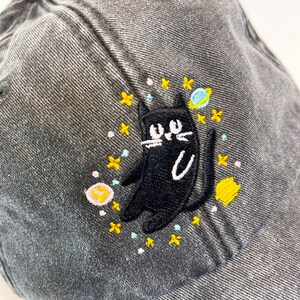 May include: A gray denim baseball cap with an embroidered black cat design. The cat is surrounded by yellow, pink, and blue stars and a small planet.