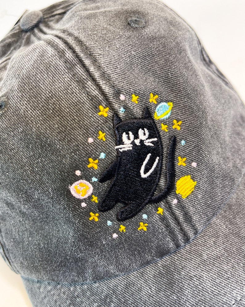 May include: A gray denim baseball cap with an embroidered black cat design. The cat is surrounded by yellow, pink, and blue stars and a small planet.
