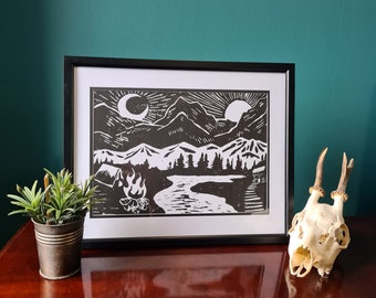 May include: Black and white framed print of a camping scene with mountains, a river, and a campfire. The print features a crescent moon and a sun. A small potted plant and a deer skull are on the table.