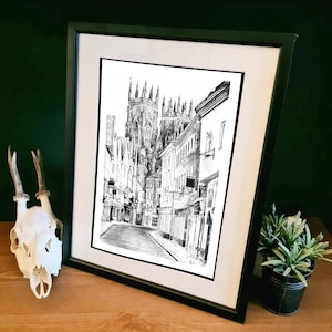 May include: Black and white drawing of a street scene in a city. The drawing features a tall church building with a pointed roof and several smaller buildings lining the street. The street is empty and the drawing is framed in a black frame.