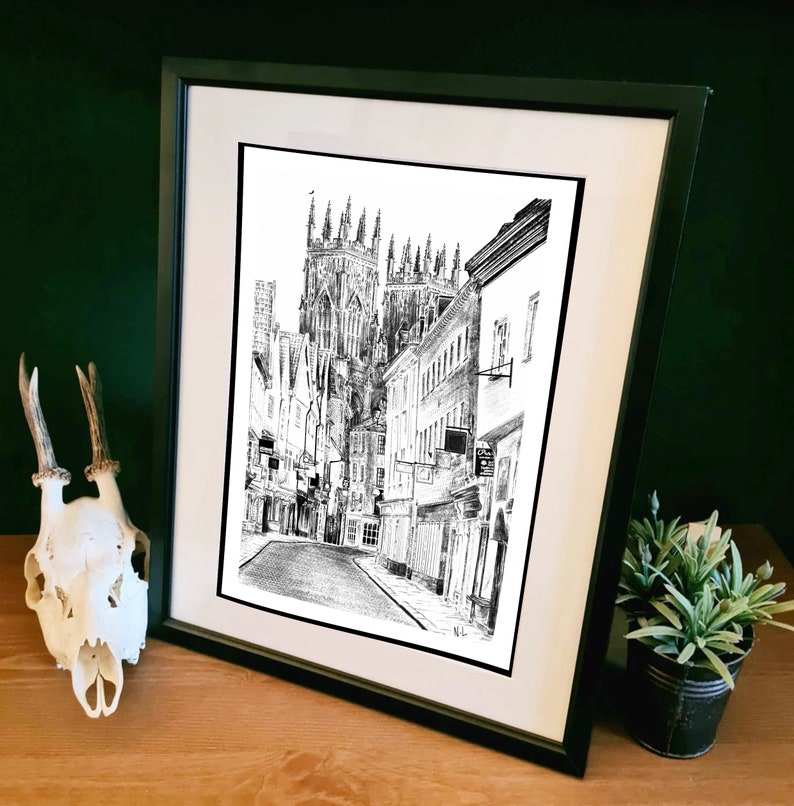 May include: Black and white drawing of a street scene in a city. The drawing features a tall church building with a pointed roof and several smaller buildings lining the street. The street is empty and the drawing is framed in a black frame.