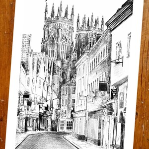 May include: A black and white pencil drawing of a street in a city with a large cathedral in the background. The street is lined with buildings and there are people walking on the sidewalk.
