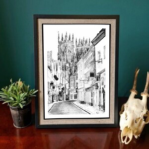 May include: Black and white ink drawing of a street scene in a city. The drawing features a tall church building with a pointed roof and several smaller buildings lining the street. The street is empty and the drawing has a vintage feel.