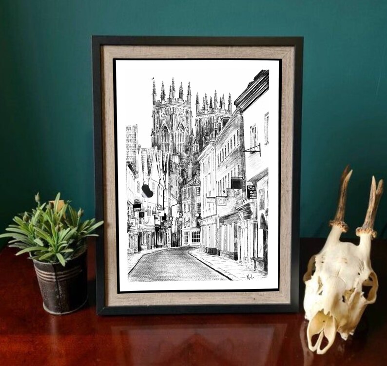 May include: Black and white ink drawing of a street scene in a city. The drawing features a tall church building with a pointed roof and several smaller buildings lining the street. The street is empty and the drawing has a vintage feel.