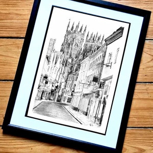 May include: A black and white ink drawing of a city street with a large cathedral in the background. The drawing is framed in a black frame with a white mat.
