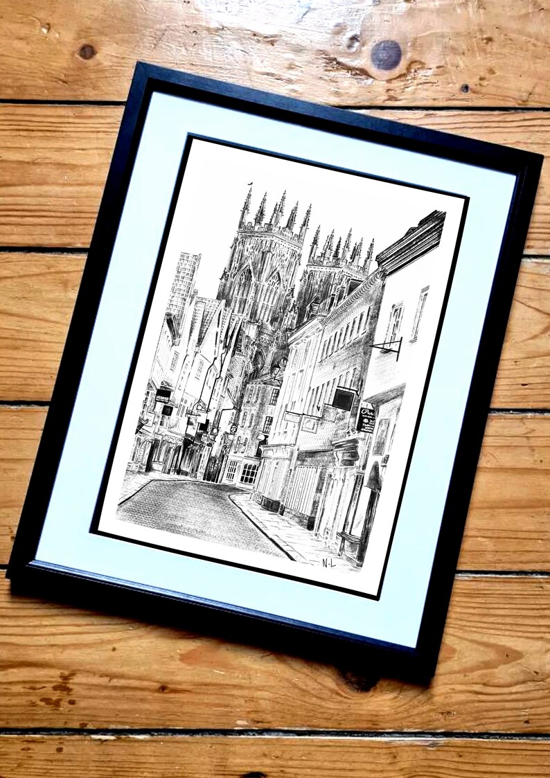 May include: A black and white ink drawing of a city street with a large cathedral in the background. The drawing is framed in a black frame with a white mat.