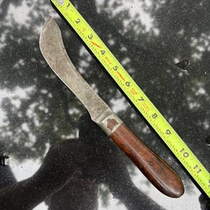 May include: A curved blade knife with a brown wooden handle and a metal bolster, positioned next to a yellow measuring tape that shows the knife has a total length of approximately 10.5 inches from the tip to the end of the handle.