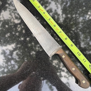 May include: A handmade chef's knife with a wooden handle and a textured, silver-coloured blade. The knife is approximately 28 cm long, as measured by a yellow tape measure. The handle is dark brown with brass rivets.