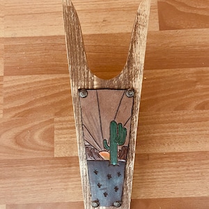 May include: Wooden boot jack with a leather inlay featuring a desert sunset scene with a cactus and a setting sun. The leather inlay is secured with metal rivets.