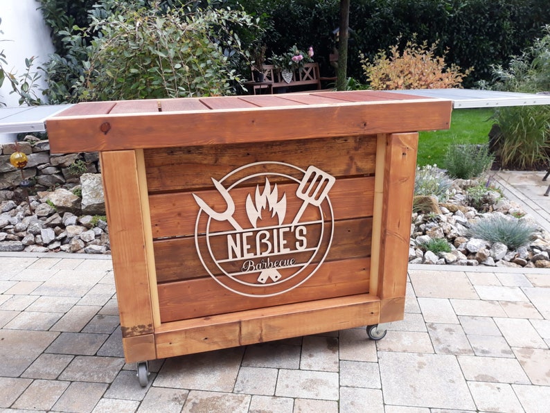 May include: A wooden mobile barbecue cart with wheels. The cart has a wooden top and a logo that says "Nebie's Barbecue" with a flame and grilling tools.
